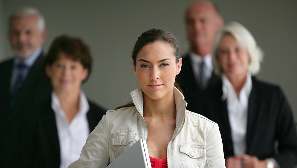 A woman in the foreground carrying a laptop with business suited people behind her.