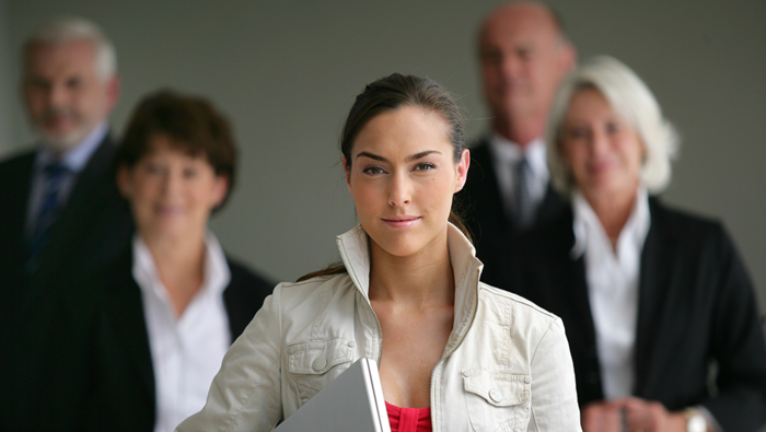A woman in the foreground carrying a laptop with business suited people behind her.